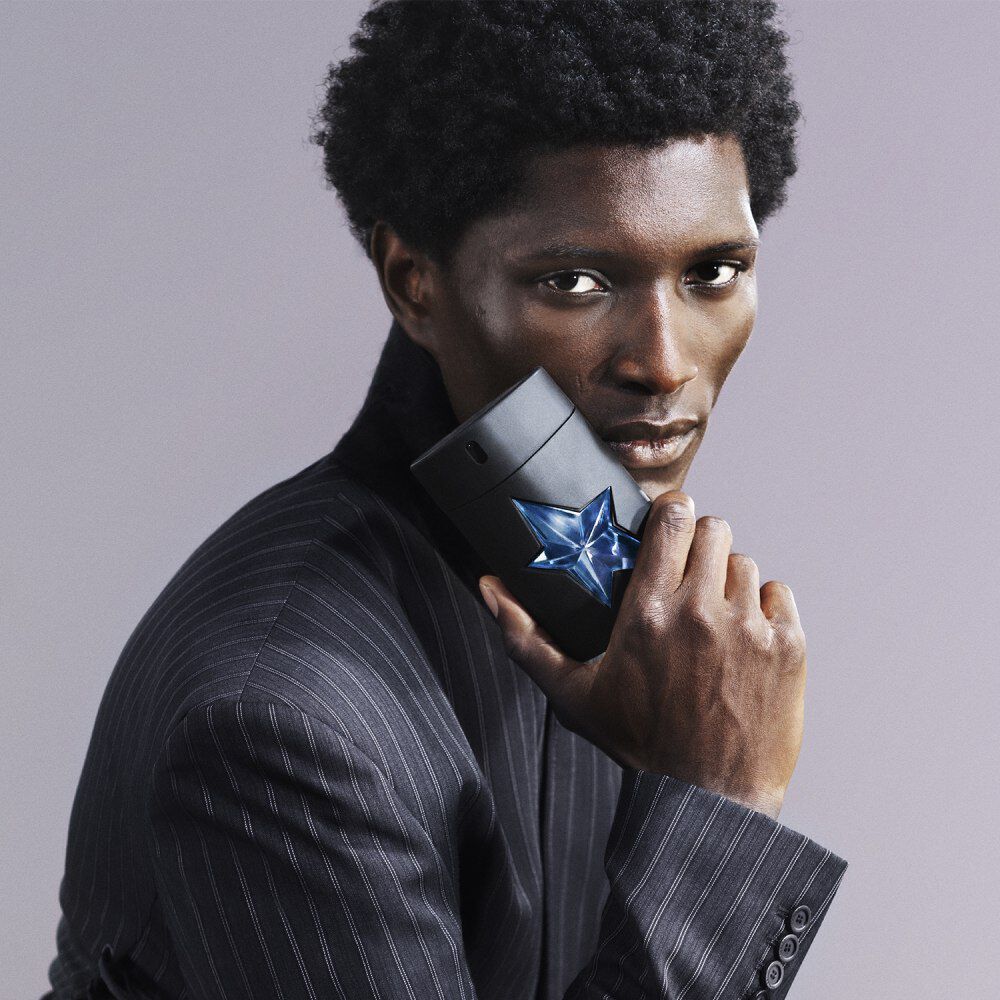 Close-up of a man in a pinstripe suit holding a matte black Mugler A*Men perfume bottle with a blue star near his cheek.