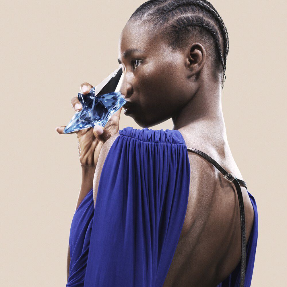 Profile of a woman in a royal blue draped dress holding a star-shaped, light blue glass Mugler Angel perfume bottle.
