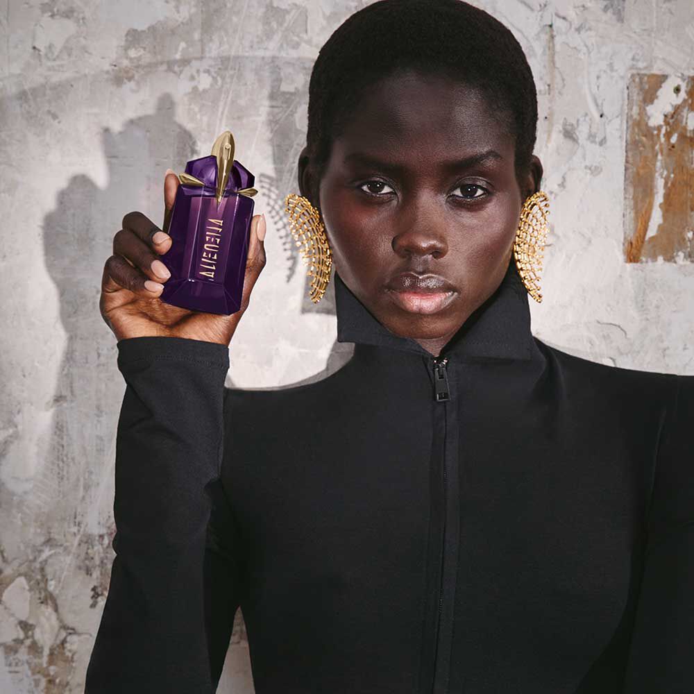 Person holding Mugler Alien purple perfume bottle against textured wall