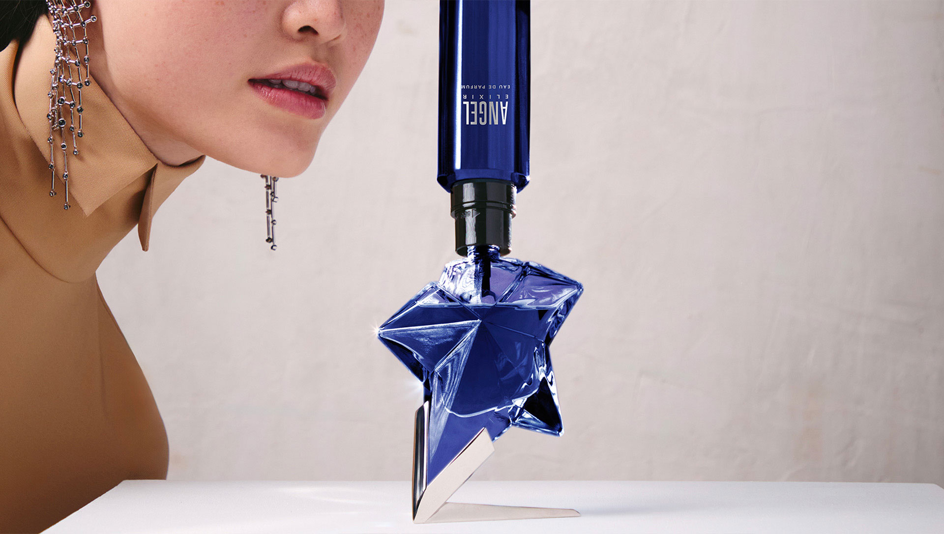 Mugler Alien Elixir blue and midnight star-shaped bottles surrounded by white flowers and yellow botanicals, showcasing key fragrance ingredients