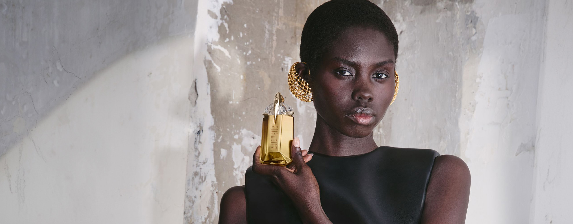 Model in black dress holding Alien perfume bottle against a textured stone wall