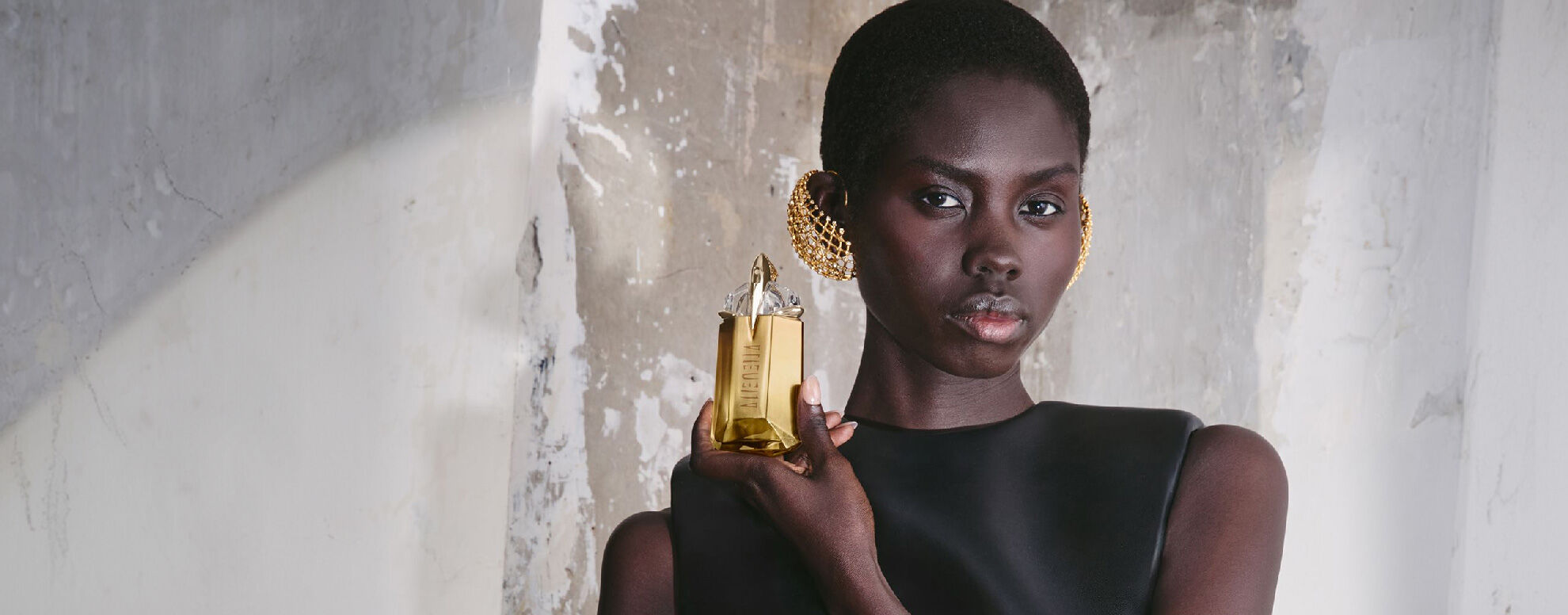 Model in black dress holding Alien perfume bottle against a textured stone wall
