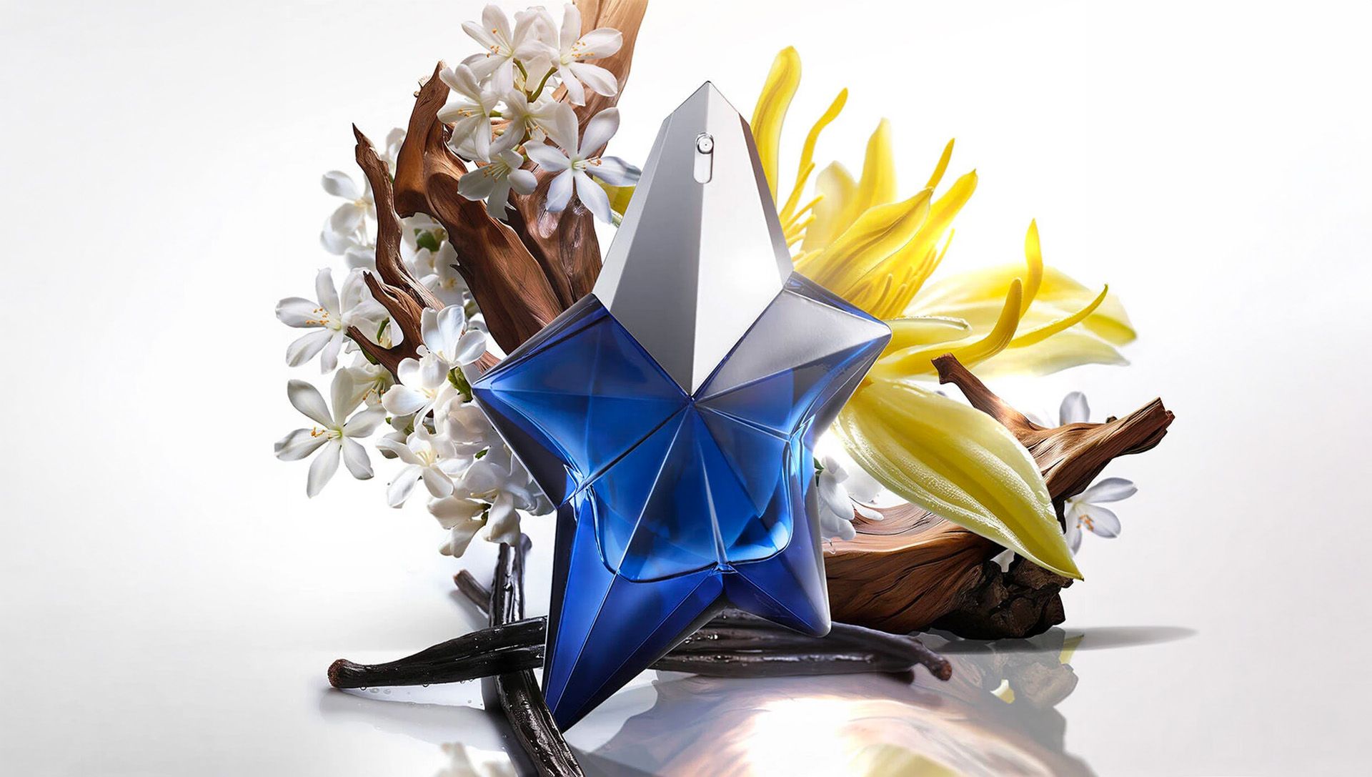 Mugler Alien Elixir blue and midnight star-shaped bottles surrounded by white flowers and yellow botanicals, showcasing key fragrance ingredients