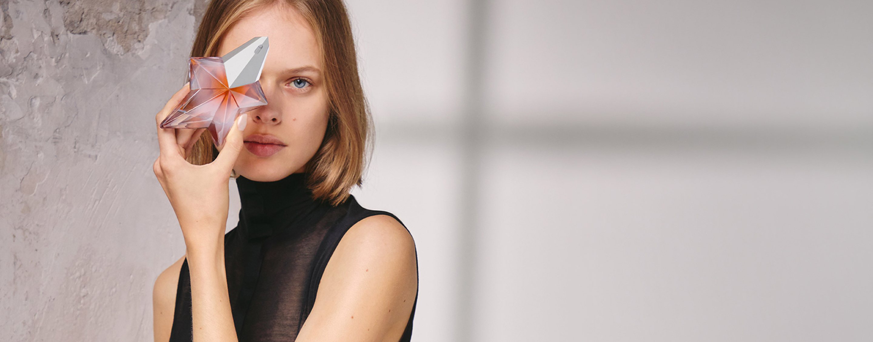 Model in a sheer black sleeveless top holding a Mugler Angel Blush perfume bottle near her face.