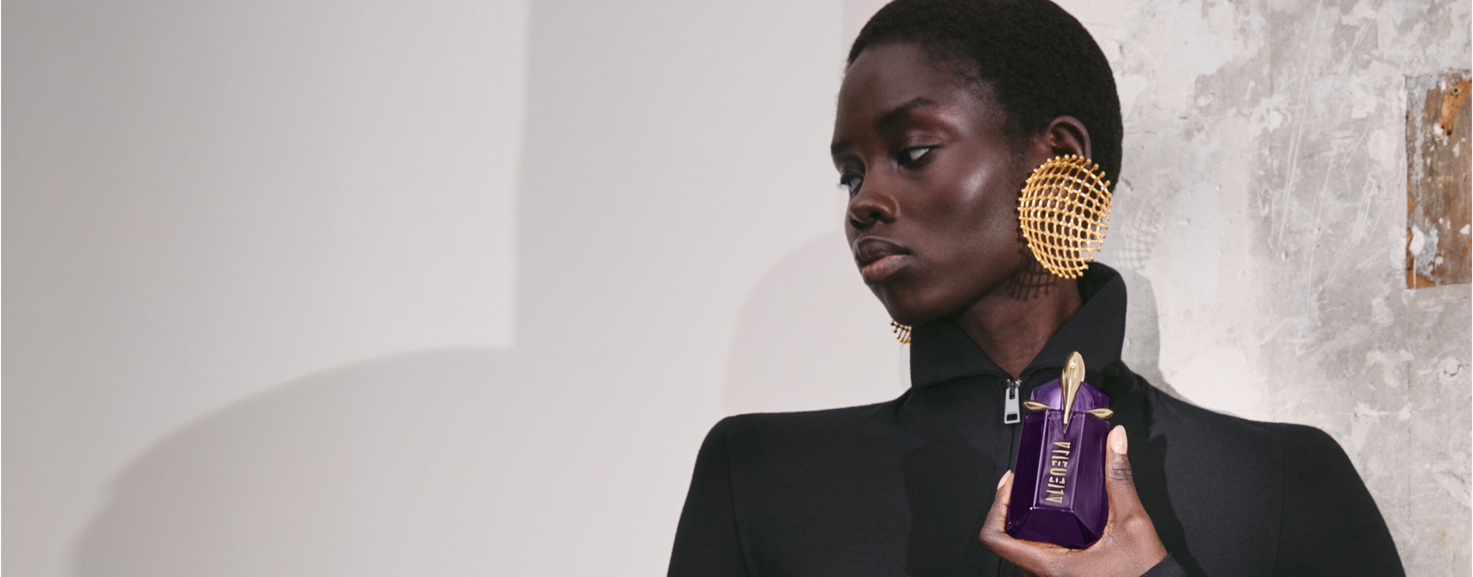 Person holding Mugler Alien purple perfume bottle against textured wall