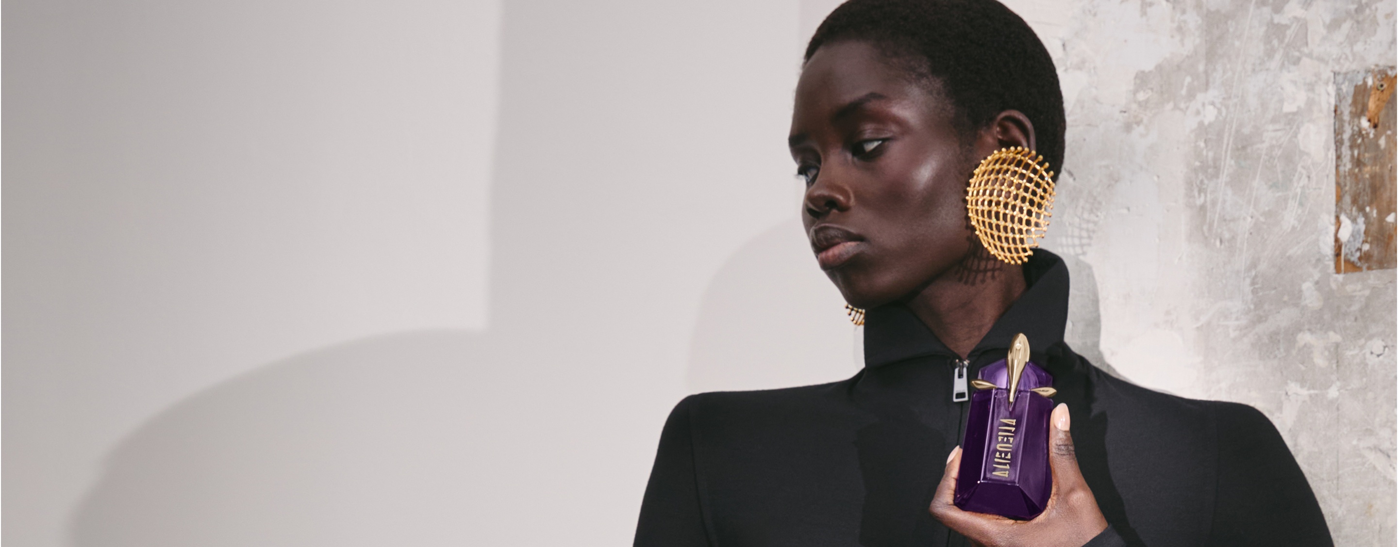 Person holding Mugler Alien purple perfume bottle against textured wall